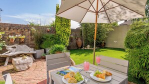 An outdoor patio with a table set for a meal, including plates of food and drinks, surrounded by greenery and a brick wall.