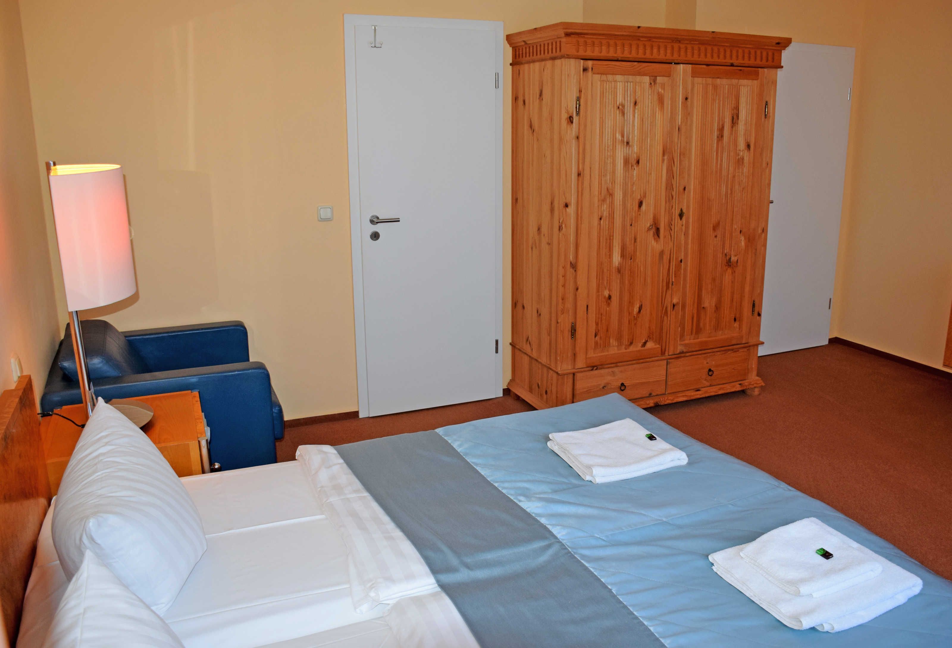 Une chambre d’hôtel avec un lit, une armoire en bois, un bureau avec une lampe et une chaise bleue.