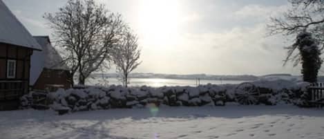A snow-covered landscape with a house, a stone wall, and a body of water in the background.