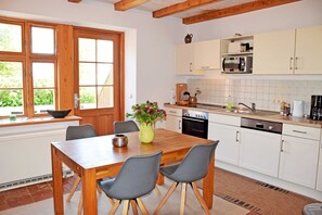 A modern kitchen with wooden cabinets, a dining table with chairs, a vase with flowers, and a window with a view of greenery.