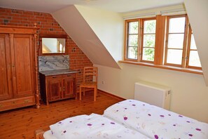 A bedroom with a wooden wardrobe, a desk, a chair, a bed with floral bedding, a radiator, and a window with wooden frames.