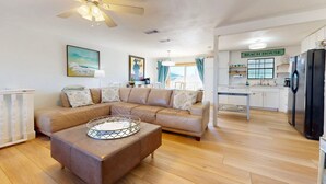 A living room with a sectional sofa, a glass coffee table, and a kitchen area with a refrigerator and a sign that reads "BEACH HOUSE". - Pet Friendly with Large Fenced Yard, Golf Cart Access (Port Aransas)