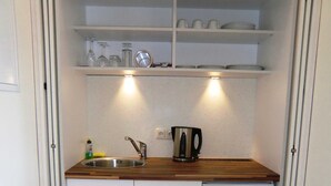 A compact kitchen with a sink, a kettle, and a shelf above containing glassware and plates. - Double room 5 - PENSION Delia Will in Ahlbeck (Heringsdorf)
