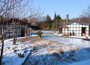 A snow-covered outdoor area with a frozen pond, bare trees, and a building with a traditional architectural style.