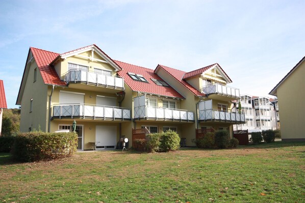 Un edificio residencial de varios pisos con balcones, techo plano y un jardín delantero con césped.