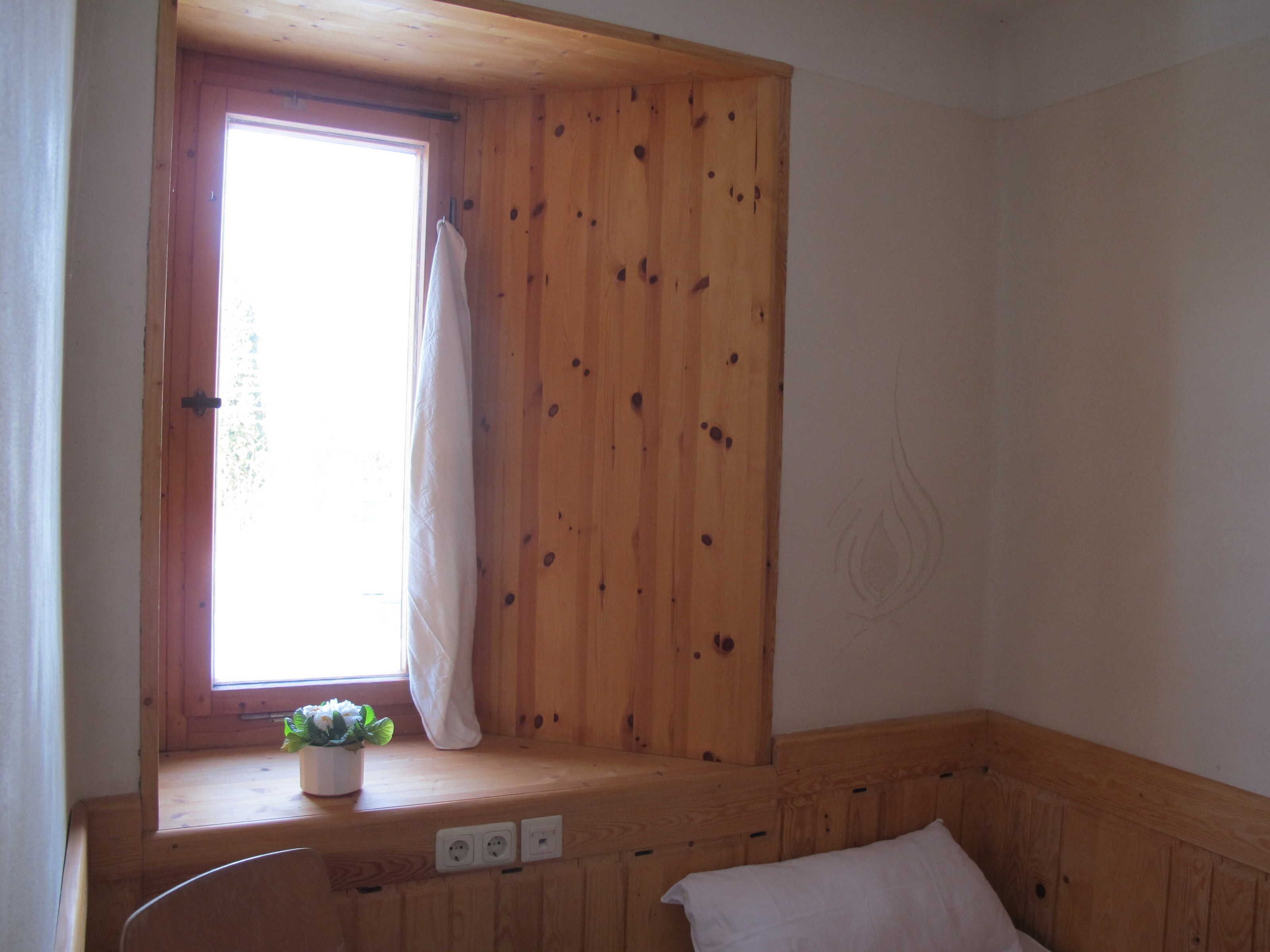 A room with wooden walls, a window with a curtain, a small plant on a ledge, and a bed with a pillow.
