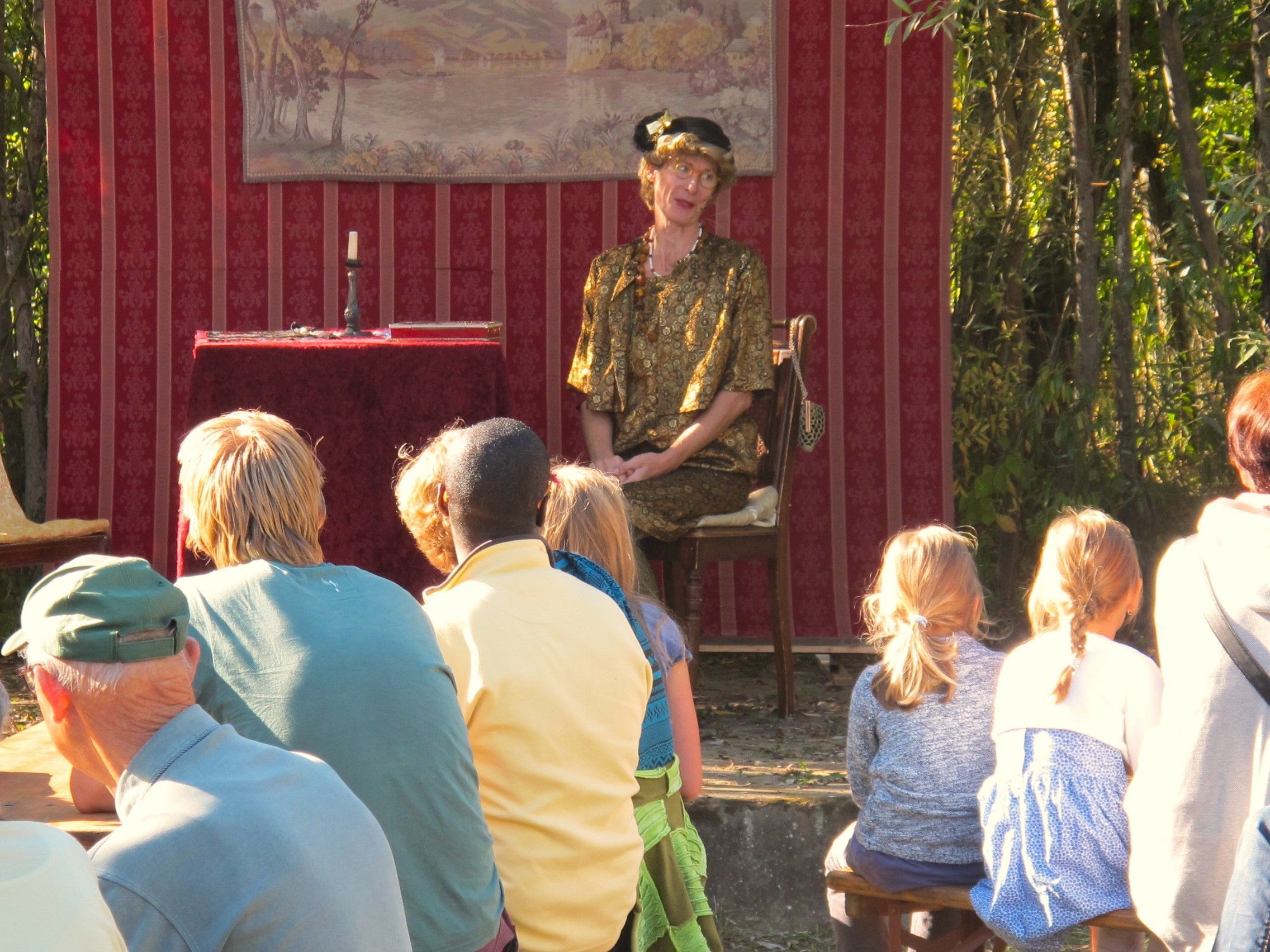 A person seated on a chair in front of a red backdrop with a painting, surrounded by an audience.