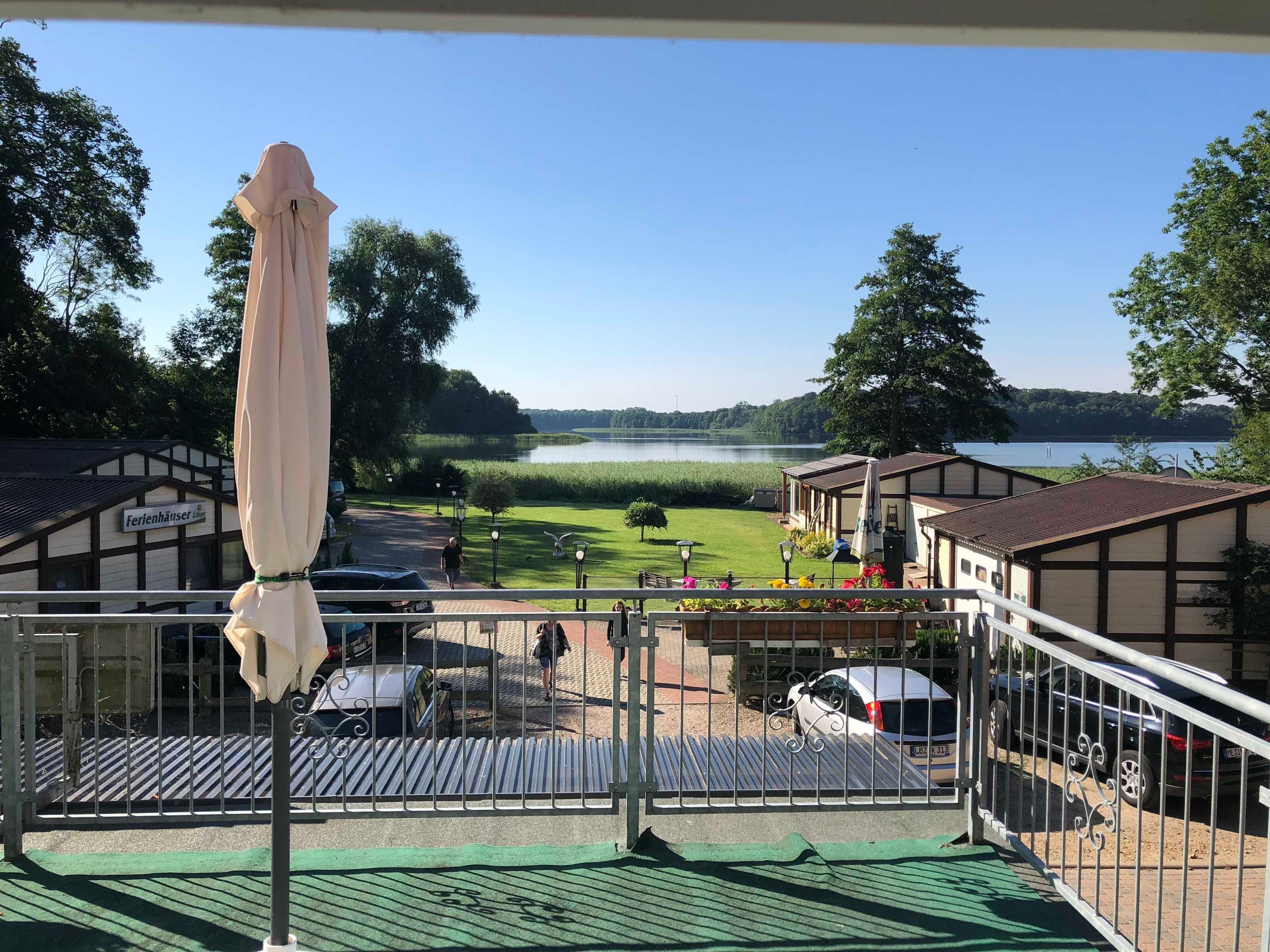 Une vue depuis un balcon donnant sur un lac, avec un bâtiment portant l’inscription « Ferlenhäuser » et un parking avec des voitures.