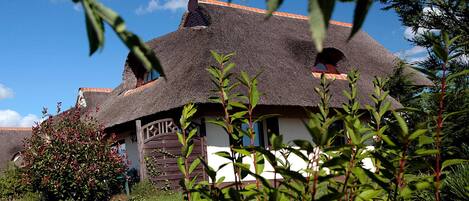 Une maison traditionnelle à toit de chaume, dotée d’une porte en bois et d’une petite fenêtre, entourée de verdure.