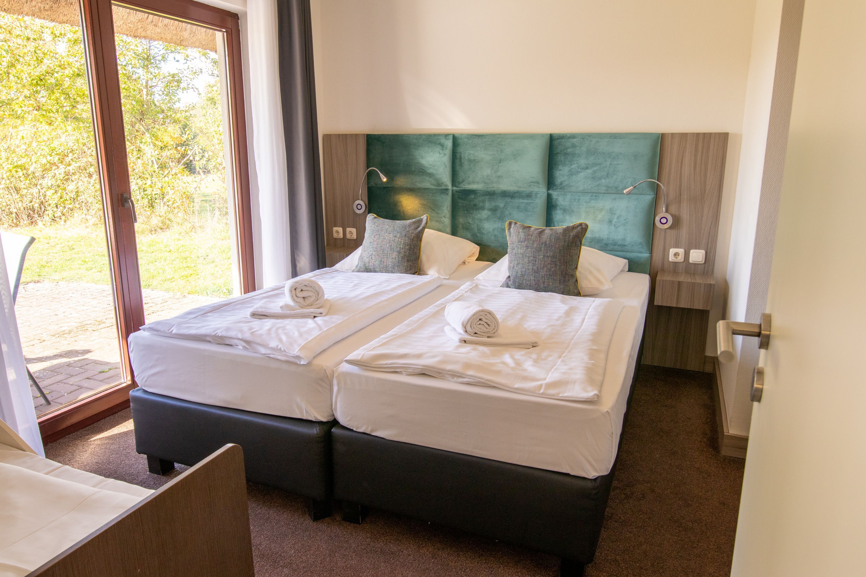 Een tweepersoonsbed met een groen hoofdeinde, twee nachtkastjes en een raam met uitzicht op groen.