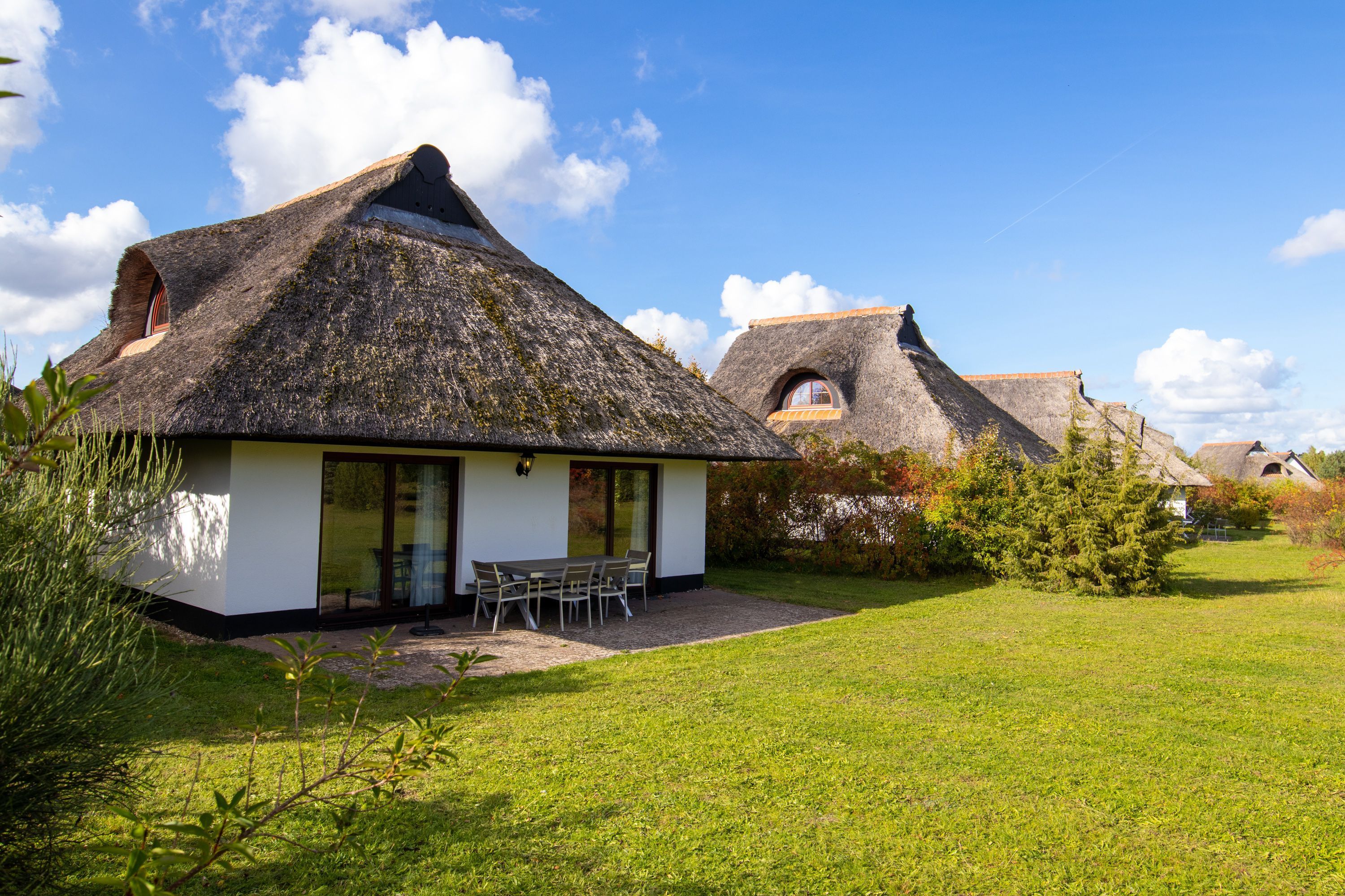 Une maison moderne avec un toit de chaume, une terrasse dotée de mobilier d’extérieur et une pelouse bien entretenue.