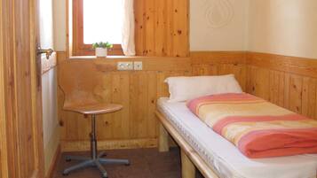 A small room with a wooden bed, a small table, a window with a curtain, and a door.