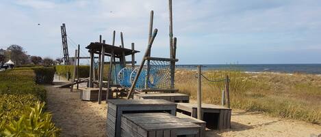 Zona arenosa de playa con estructuras de madera, incluyendo un refugio de madera y un área de juegos con mosquitero.