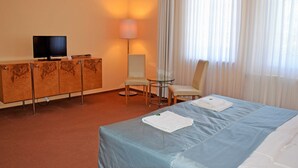 Quarto de hotel com uma cama, uma televisão sobre um móvel de madeira, uma escrivaninha com uma lâmpada e duas cadeiras.