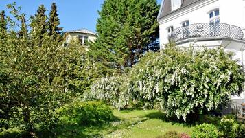 Ein üppiger Garten mit verschiedenen Bäumen und Sträuchern, ein weißes Gebäude mit Balkon und ein klarer, blauer Himmel.