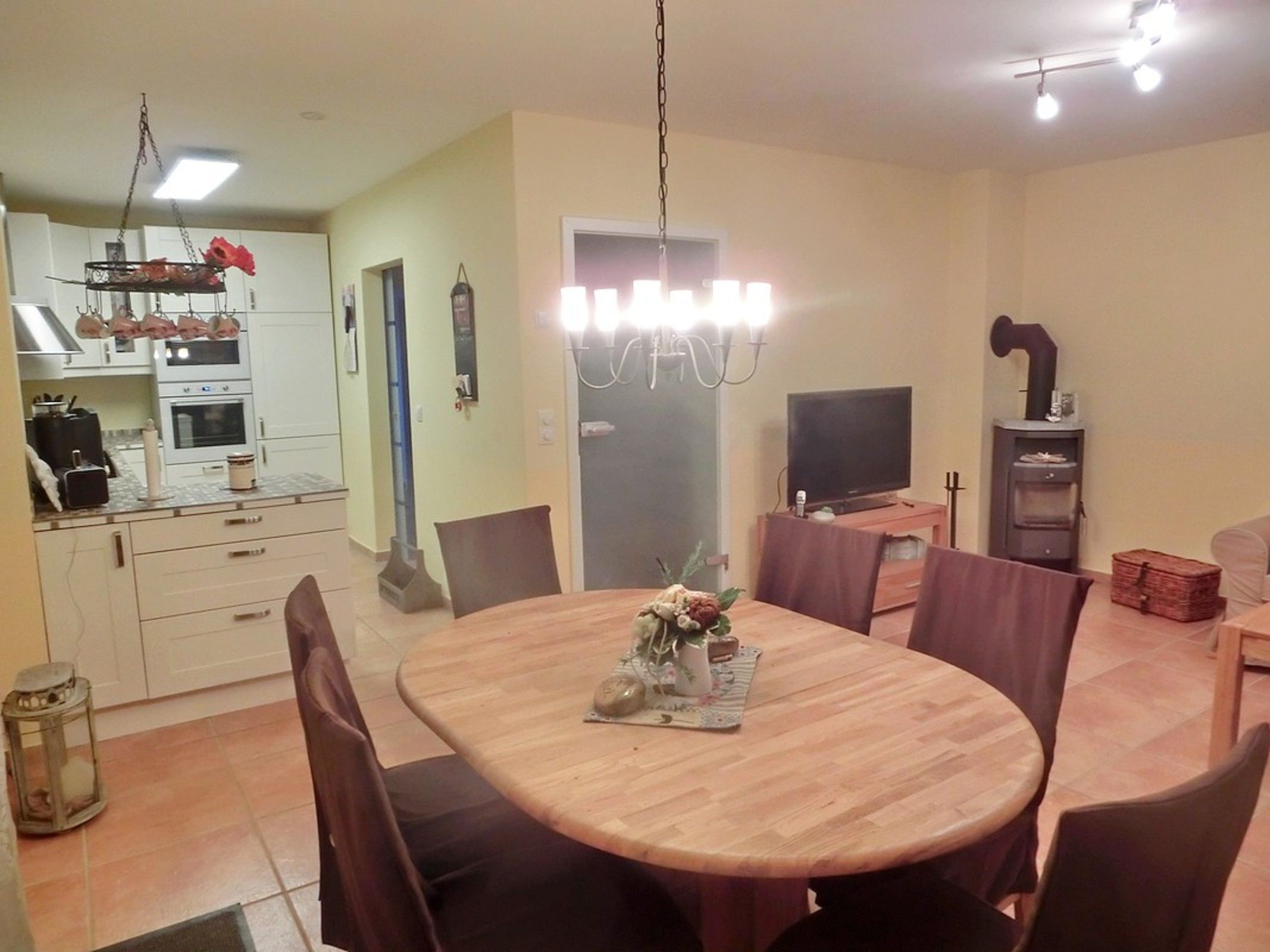 A dining area with a round wooden table, six chairs, a chandelier, a television, and a wood-burning stove.