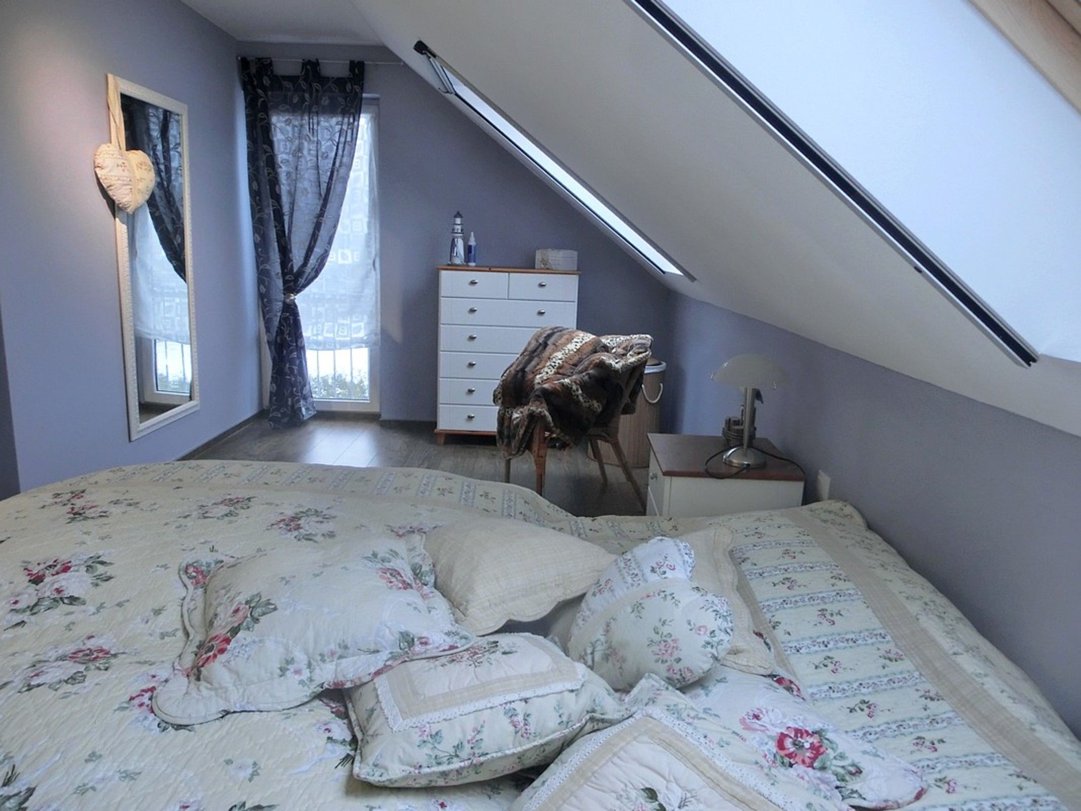 Et soveværelse med skråvæg, seng med blomstret sengetøj, kommode, lampe og et vindue med gardiner.