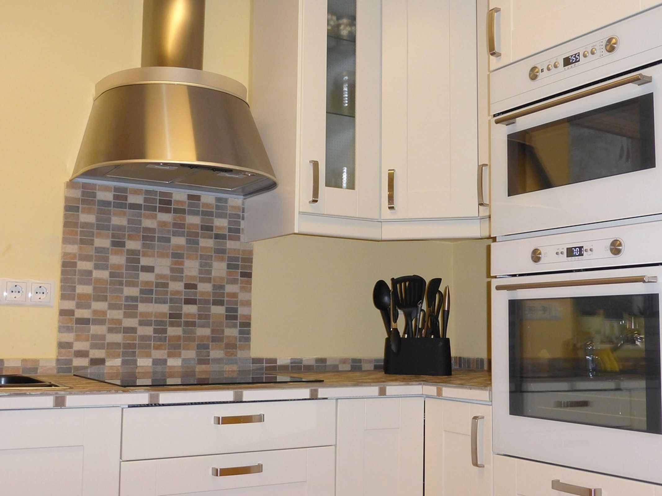 A kitchen with white cabinets, a stainless steel range hood, a tiled backsplash, and two ovens.