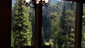 A dining area with a table and chairs, large windows with a view of a forest, and a chandelier. - Ski Heaven,  Ski in, Ski out Big Sky Mtn Village Home with Hot Tub (Big Sky)