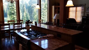 A kitchen with a central island, wooden chairs, a stove, and a view of a forest through large windows. - Ski Heaven,  Ski in, Ski out Big Sky Mtn Village Home with Hot Tub (Big Sky)