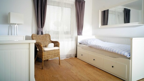 Une chambre à coucher avec un lit blanc, une chaise en osier, une commode blanche, un miroir et une fenêtre avec des rideaux.