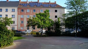 Un immeuble à plusieurs étages, doté de panneaux solaires sur le toit, d’une cour pavée et de voitures garées.