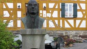 Busto de Franz Daniel Pastorius sobre um pedestal com uma placa, em frente a um edifício com fachada amarela e branca marcante.