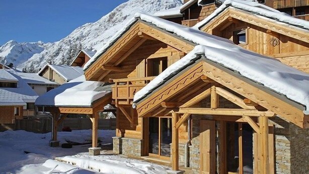 Una hilera de cabañas de madera con tejados nevados, con montañas nevadas al fondo.