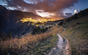 Un village de montagne au crépuscule, avec un sentier sinueux menant à un hameau illuminé.