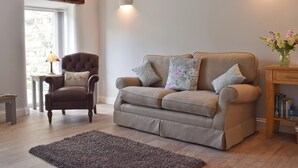 Un salon avec un fauteuil marron, un canapé gris et une table basse en bois. Il y a une fenêtre avec des rideaux, un tapis sur le sol et un vase avec des fleurs sur une table d’appoint.