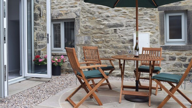 Un espace extérieur aménagé avec des sièges, un mur en pierre, des chaises en bois, une table et un parasol vert.