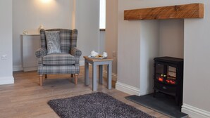 Un salon avec un fauteuil à carreaux, une petite table sur laquelle se trouve un plateau de biscuits, une cheminée et un tableau accroché au mur.