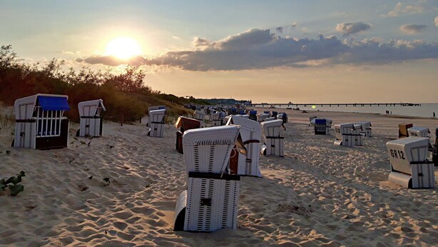En strand med rader av vita strandstolar och parasoller, en pir i bakgrunden och solen som går ner över horisonten.