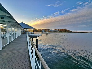 Eine Pier am Wasser mit einem überdachten Sitzbereich und Blick auf das Wasser sowie entfernte Gebäude.