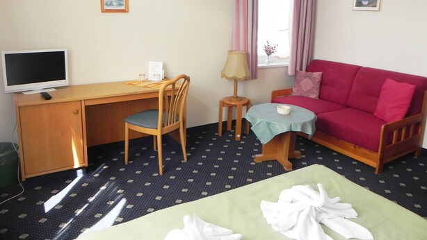Une chambre équipée d’un bureau, d’une chaise, d’une télévision, d’un canapé rouge, d’une petite table, d’une lampe et d’un lit avec des serviettes.