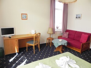 Ein Zimmer mit Schreibtisch, Stuhl, Fernseher, rotem Sofa, kleinem Tisch, Lampe und einem Bett mit Handtüchern.