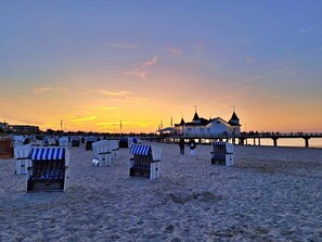 Une plage avec des chaises longues et des parasols, une jetée et un bâtiment doté d’une tour.