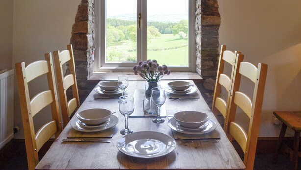 A dining table set for four with chairs, a vase with flowers, and a window with a view of a green landscape. - Mountain View-UK11825 (Sarnau, near Brecon)