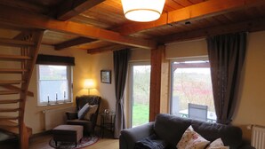 A cozy living room with a wooden ceiling, a sofa with cushions, a chair, a small table with a lamp, and a view through a window.