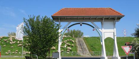 A rural entrance with a white archway, a red-tiled roof, and a grassy hill with sheep.