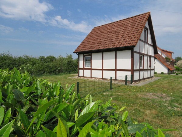 A traditional half-timbered house with a sloped roof, surrounded by lush greenery and a clear sky.