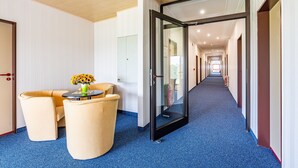 Un couloir d’hôtel avec une moquette bleue, une porte en verre, un canapé et une petite table ornée de fleurs.