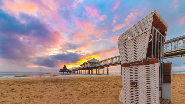 Une plage avec une jetée en bois menant à un bâtiment doté d’un toit triangulaire.