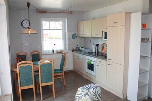 A compact kitchen with light wooden cabinets, a microwave, and a sink. There is a dining table with chairs and a window with a view outside.
