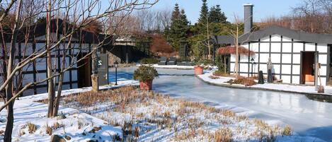 A snow-covered landscape with a frozen pond, bare trees, and traditional buildings with large windows.