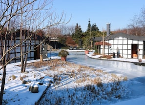 A snow-covered landscape with a frozen pond, bare trees, and traditional buildings with large windows.