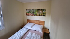 Un dormitorio con cama de madera, ropa de cama blanca, una mesita de noche, un cuadro de un bosque y una ventana con puerta.