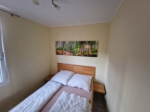 Camera da letto con un letto in legno, biancheria bianca, un comodino, un quadro che raffigura una foresta e una finestra con una porticina.