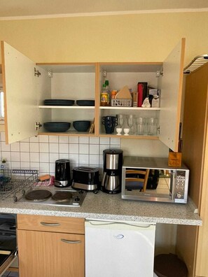 Une cuisine compacte équipée d’un four à micro-ondes, d’un grille-pain et d’une machine à café intégrés. Des étagères au-dessus du plan de travail servent à ranger la vaisselle et les verres.