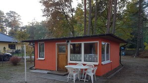 Un pequeño edificio de una sola planta con fachada roja y detalles blancos, que presenta una puerta de madera y ventanas amplias. Cuenta con un pequeño patio con una mesa y sillas.